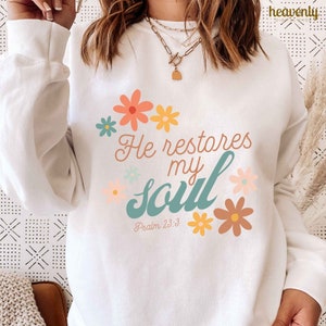 Puede incluir: Sudadera blanca con un dise&ntilde;o floral en azul verdoso, amarillo y rosa. El texto "He restores my soul" est&aacute; escrito en una fuente de letra cursiva azul verdoso con las palabras "Psalm 23:3" debajo.