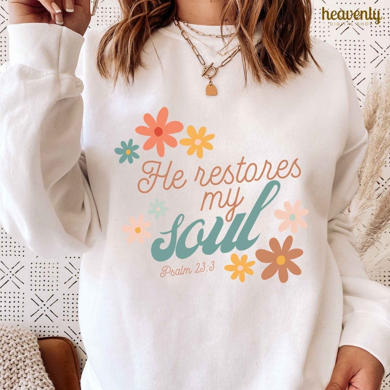 Puede incluir: Sudadera blanca con un dise&ntilde;o floral en azul verdoso, amarillo y rosa. El texto "He restores my soul" est&aacute; escrito en una fuente de letra cursiva azul verdoso con las palabras "Psalm 23:3" debajo.