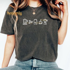 May include: A dark gray t-shirt with a white graphic of coffee brewing equipment and coffee beans. The text "heavenly" is in yellow above the graphic.