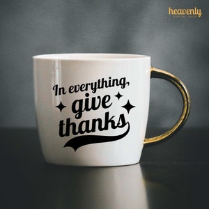 May include: White ceramic mug with a gold handle. The mug has a black graphic that says "In everything, give thanks" with two small stars on either side of the word "give".