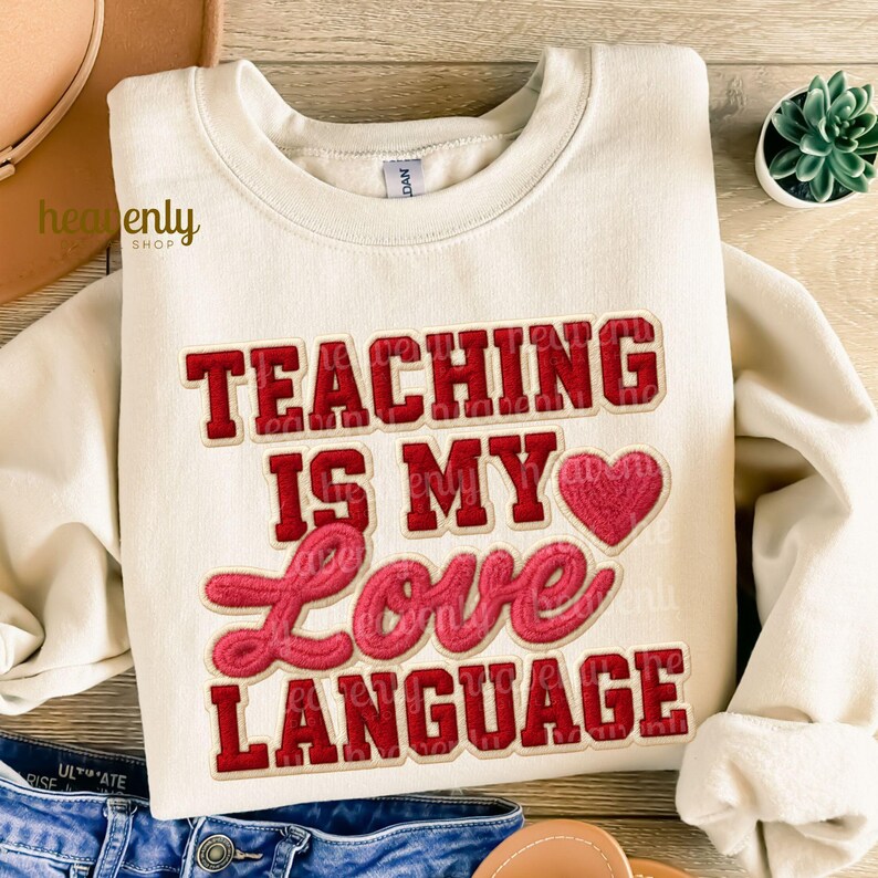 K&ouml;nnte beinhalten: Cremefarbenes Sweatshirt mit dem Schriftzug "TEACHING IS MY LOVE LANGUAGE" in roter und rosa Schrift. Das Design beinhaltet ein rotes Herz. Das Sweatshirt ist auf einer Holzoberfl&auml;che gefaltet.