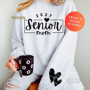 May include: A light gray sweatshirt with the text "2027 Senior Mom" in black. The sleeve features two black hearts. A black mug with white flower designs is held in the hand. The sweatshirt design is for front and sleeve designs.
