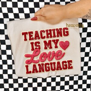 K&ouml;nnte beinhalten: Cremefarbenes T-Shirt mit dem Text "TEACHING IS MY LOVE LANGUAGE" in roter und rosa Schrift. Eine rote Herzgrafik befindet sich zwischen "MY" und "LOVE". Das Shirt wird vor einem schwarz-wei&szlig; karierten Hintergrund gehalten.