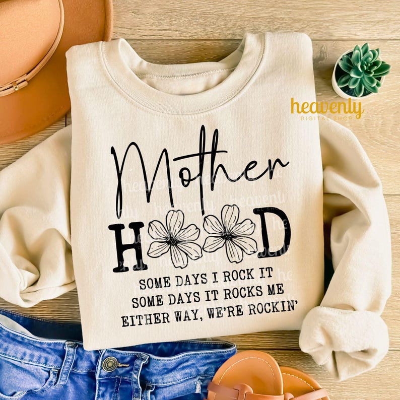May include: A beige sweatshirt with the text "Motherhood" in black lettering with two black flowers on either side of the "H". The text below reads "Some days I rock it, some days it rocks me, either way, we're rockin'."