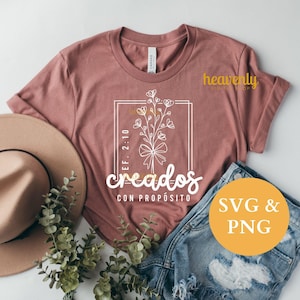 May include: A dusty rose t-shirt with a floral design inside a white rectangular frame. The text "creados con propósito" is below the design. A tan hat and denim shorts are next to the shirt. A yellow circle with "SVG & PNG" is on the right.