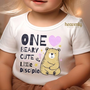 May include: White onesie with a pink heart, a brown bear, and the text "ONE BEARY CUTE Little Disciple".