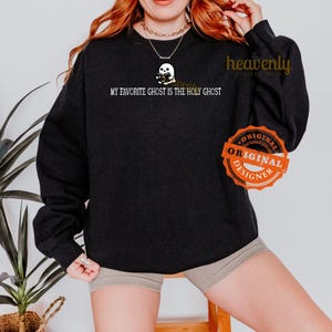 K&ouml;nnte beinhalten: Schwarzes Sweatshirt mit dem Text "My favorite ghost is the Holy Ghost" und einem Cartoon-Geist, der eine Kamera h&auml;lt. Das Sweatshirt hat ein wei&szlig;es "Original Designer"-Label.