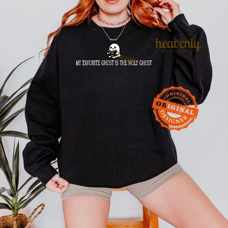 K&ouml;nnte beinhalten: Schwarzes Sweatshirt mit dem Text "My favorite ghost is the Holy Ghost" und einem Cartoon-Geist, der eine Kamera h&auml;lt. Das Sweatshirt hat ein wei&szlig;es "Original Designer"-Label.