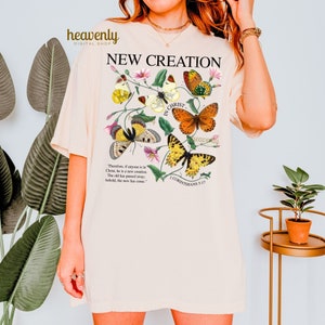 May include: A beige t-shirt with a floral design and butterflies. The text "NEW CREATION" is printed in black at the top of the shirt. The text "IN CHRIST" is printed in brown with a floral design. The text "Therefore, if anyone is in Christ, he is a new creation. The old has passed away; behold, the new has come." is printed in black. The text "2 CORINTHIANS 5:17" is printed in black at the bottom of the shirt.