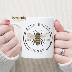 May include: White ceramic mug featuring a detailed illustration of a bee with the text "KIND WORDS ARE LIKE HONEY" and "PROVERBS 16:24". The mug has a curved handle and is held by two hands.