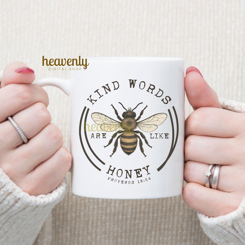 May include: White ceramic mug featuring a detailed illustration of a bee with the text "KIND WORDS ARE LIKE HONEY" and "PROVERBS 16:24". The mug has a curved handle and is held by two hands.