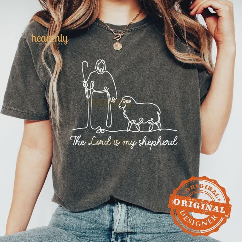 Pu&ograve; includere: T-shirt grigio scuro con un disegno a tratto bianco di un pastore e una pecora, e la scritta "The Lord is my shepherd". La maglietta ha girocollo e maniche corte. Il disegno &egrave; centrato.