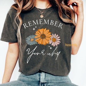 K&ouml;nnte beinhalten: Ein dunkelgraues T-Shirt mit dem Text "REMEMBER your why" in Wei&szlig;. Der Text ist von drei stilisierten Blumen in Orange, Rosa und Blau umgeben.