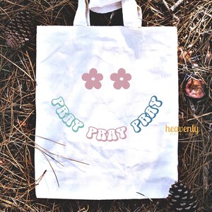 May include: A white canvas tote bag with a smiling face design. The design includes two pink flower shapes for eyes and the word "PRAY" in a curved, retro font, in shades of blue and pink. The bag is sitting on a bed of pine needles.