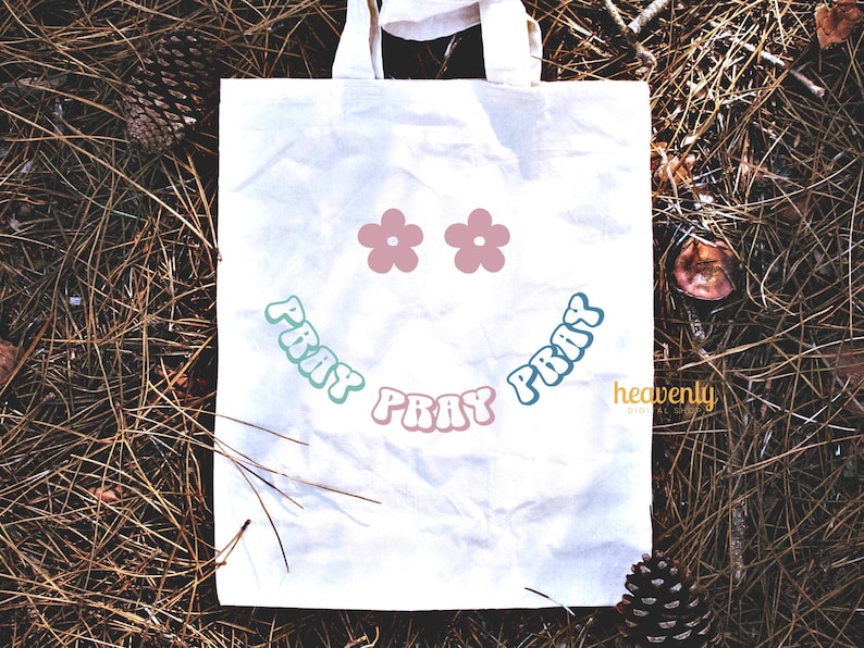 May include: A white canvas tote bag with a smiling face design. The design includes two pink flower shapes for eyes and the word "PRAY" in a curved, retro font, in shades of blue and pink. The bag is sitting on a bed of pine needles.