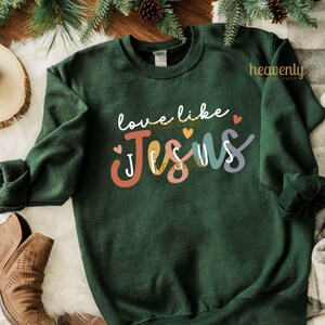 May include: A dark green sweatshirt with the words "love like Jesus" in white script and the word "Jesus" in colorful block letters. The design includes small hearts and is set against a cozy background.