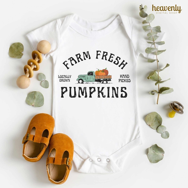 May include: White baby bodysuit featuring the text "Farm Fresh Pumpkins" and a graphic of a vintage green truck loaded with pumpkins. The image includes baby shoes, a wooden rattle, and foliage.