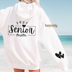 May include: White hoodie with the text "Senior mom" in a black rectangular frame, with the year "2027" above it. Two black heart graphics are on the sleeve. The words "heavenly DIGITAL SHOP" are printed on the sleeve.