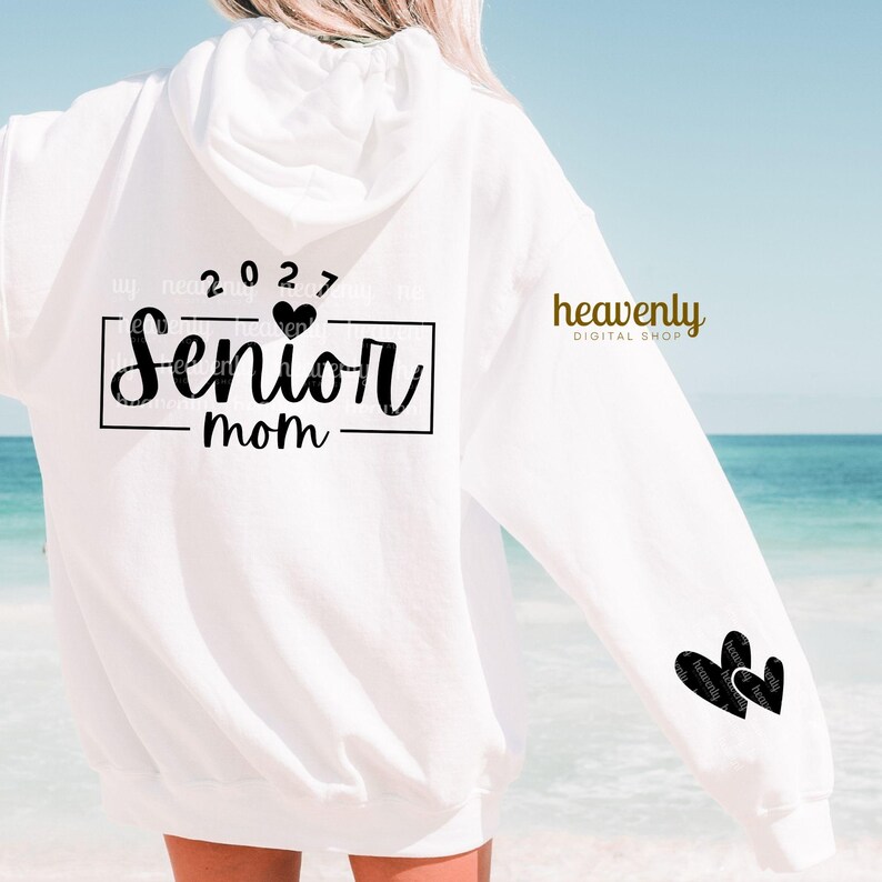 May include: White hoodie with the text "Senior mom" in a black rectangular frame, with the year "2027" above it. Two black heart graphics are on the sleeve. The words "heavenly DIGITAL SHOP" are printed on the sleeve.