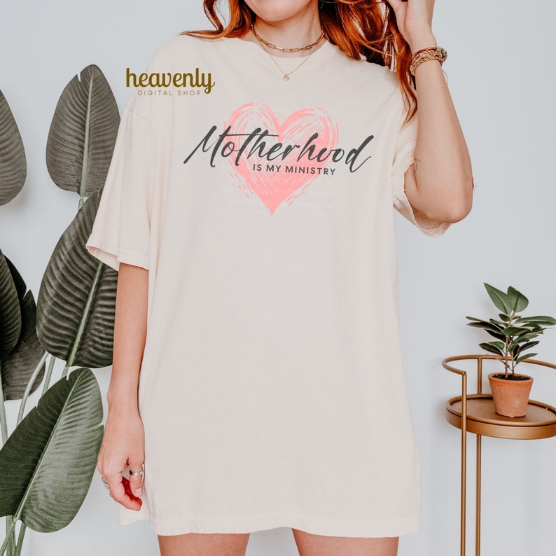 Op de afbeelding: Een wit T-shirt met een roze hart en de tekst "Motherhood is my ministry".
