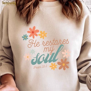 Puede incluir: Una sudadera beige con un dise&ntilde;o floral y el texto "He restores my soul" en turquesa y blanco. El texto "Psalm 23:3" tambi&eacute;n est&aacute; incluido en el dise&ntilde;o.