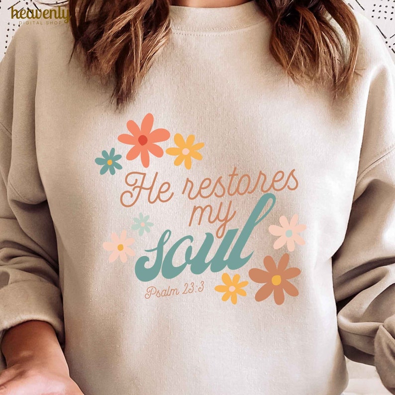 Puede incluir: Una sudadera beige con un dise&ntilde;o floral y el texto "He restores my soul" en turquesa y blanco. El texto "Psalm 23:3" tambi&eacute;n est&aacute; incluido en el dise&ntilde;o.