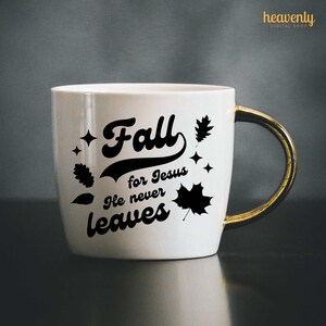 Op de afbeelding: Een witte keramische mok met een gouden handvat. De mok heeft een zwart ontwerp met de tekst "Fall for Jesus He never leaves" met gestileerde bladeren.