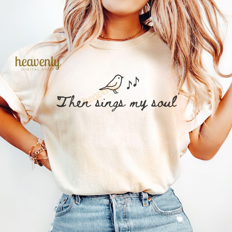 May include: Cream-colored t-shirt with a black bird and musical note graphic, and the text "Then sings my soul." The shirt is made of a soft material and is suitable for casual wear.