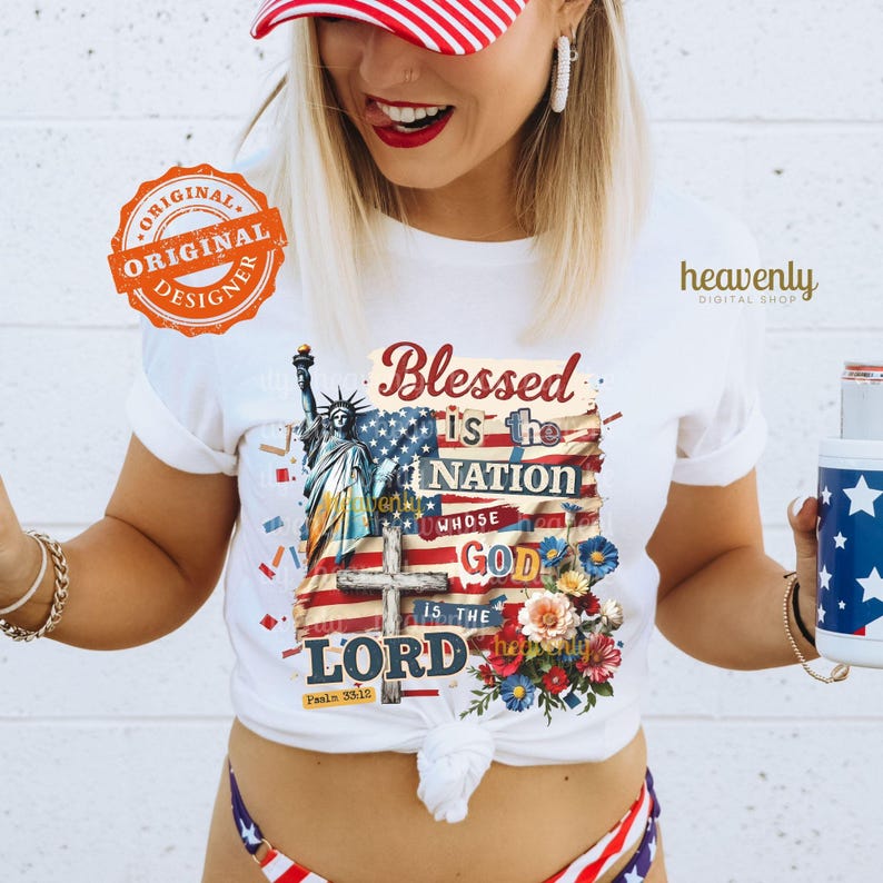 Puede incluir: Camiseta blanca con un dise&ntilde;o patri&oacute;tico que presenta la Estatua de la Libertad, una bandera estadounidense y el texto "Blessed is the Nation Whose God is the Lord". La camiseta tambi&eacute;n tiene un arreglo floral y el texto "Psalms 33:12."