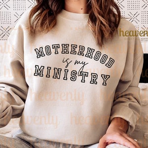 Pu&ograve; includere: Una felpa beige con il testo "MOTHERHOOD is my MINISTRY" in lettere nere.