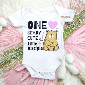 May include: White baby onesie with a pink heart, a brown bear, and the text "ONE BEARY CUTE LITTLE DISCIPLE".