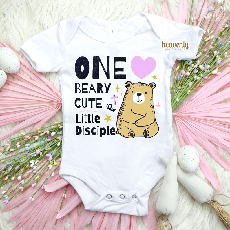 May include: White baby onesie with a pink heart, a brown bear, and the text "ONE BEARY CUTE LITTLE DISCIPLE".