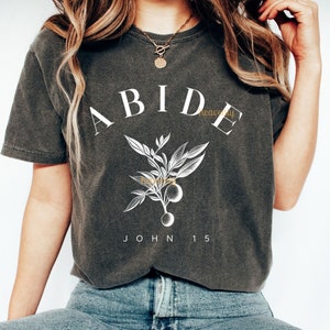 May include: A dark gray t-shirt with a white graphic design featuring the word "ABIDE" in large letters, a stylized branch with leaves and fruit, and the text "JOHN 15".