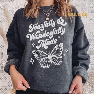 Pu&ograve; includere: Felpa grigio scuro con la scritta "Fearfully & Wonderfully Made" e una grafica a farfalla bianca. Il design include quattro piccoli diamanti. La felpa ha maniche lunghe e girocollo.