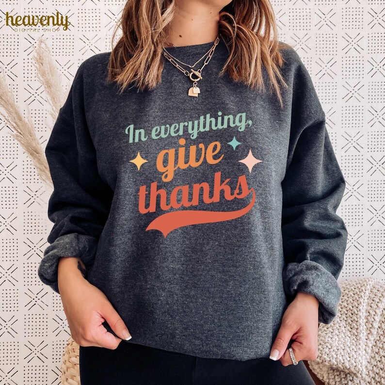 May include: A dark gray sweatshirt with the text "In everything, give thanks" in a retro font with colorful accents. The text is in a curved shape with a red, orange, and yellow color scheme. There are small yellow stars on either side of the text.