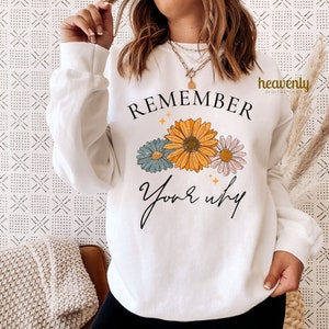 K&ouml;nnte beinhalten: Wei&szlig;er Pullover mit dem Text "REMEMBER your why" in schwarzer Schrift. Der Text ist von drei stilisierten Blumen in Gelb-, Blau- und Rosat&ouml;nen umgeben.