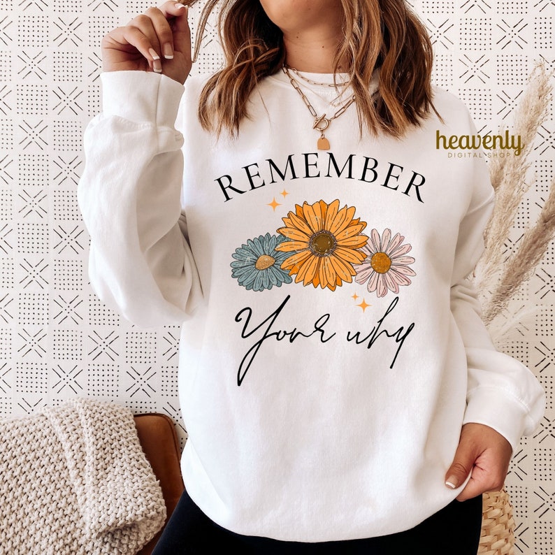 K&ouml;nnte beinhalten: Wei&szlig;er Pullover mit dem Text "REMEMBER your why" in schwarzer Schrift. Der Text ist von drei stilisierten Blumen in Gelb-, Blau- und Rosat&ouml;nen umgeben.