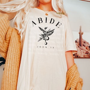 May include: A white t-shirt with a black graphic design featuring a branch with leaves and berries. The text "ABIDE" is written in a large font above the branch and the text "JOHN 15" is written below the branch.