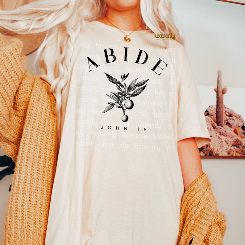 May include: A white t-shirt with a black graphic design featuring a branch with leaves and berries. The text "ABIDE" is written in a large font above the branch and the text "JOHN 15" is written below the branch.