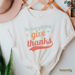 May include: A white t-shirt with the text "In everything, give thanks" in a colorful retro font. The text is in a red, orange, yellow, green, and blue color scheme. The shirt is on a hanger and has a knot tied in the bottom.