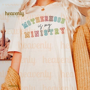 Pu&ograve; includere: Una maglietta bianca con il testo "MOTHERHOOD is my MINISTRY" in colori arcobaleno. Il testo &egrave; contornato di nero.