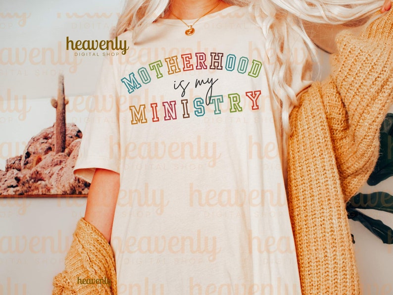 Pu&ograve; includere: Una maglietta bianca con il testo "MOTHERHOOD is my MINISTRY" in colori arcobaleno. Il testo &egrave; contornato di nero.