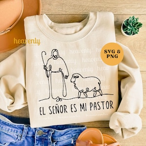Op de afbeelding: Een beige sweatshirt met een zwarte lijntekening van een herder en een schaap, met de tekst "EL SEÑOR ES MI PASTOR" eronder. Het woord "heavenly" staat herhaaldelijk op de achtergrond gedrukt. Een oranje cirkel met "SVG & PNG" staat rechtsboven.