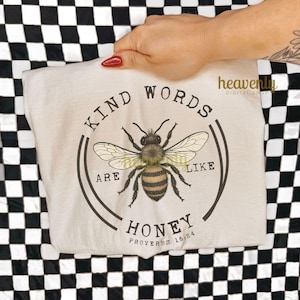 May include: A cream-colored t-shirt featuring a detailed illustration of a bee with the text "KIND WORDS ARE LIKE HONEY" in a circular design. The shirt also includes the Bible verse Proverbs 16:24.