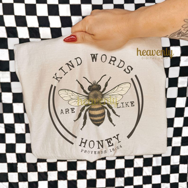 May include: A cream-colored t-shirt featuring a detailed illustration of a bee with the text "KIND WORDS ARE LIKE HONEY" in a circular design. The shirt also includes the Bible verse Proverbs 16:24.