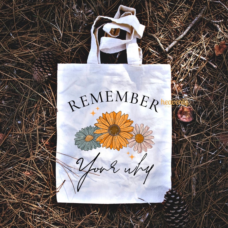 K&ouml;nnte beinhalten: Eine wei&szlig;e Canvas-Tragetasche mit dem Text "REMEMBER your why" in schwarzer Schrift. Der Text ist von drei stilisierten Blumen in Orange-, Gr&uuml;n- und Rosat&ouml;nen umgeben.