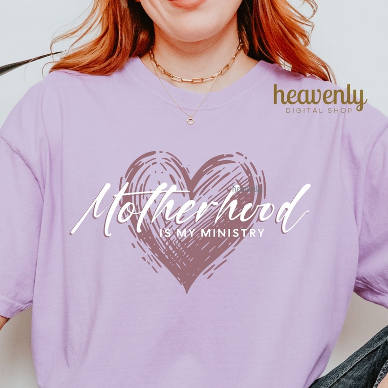 Op de afbeelding: Een paars T-shirt met een roze hart en de tekst "Motherhood is my ministry".