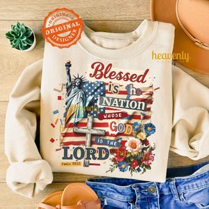 Puede incluir: Sudadera color crema con un dise&ntilde;o gr&aacute;fico que presenta la Estatua de la Libertad, una bandera estadounidense, una cruz y detalles florales. El texto dice "Blessed is the Nation Whose God is the Lord" con Salmo 33:12. Un logotipo circular "Original Designer" es visible.