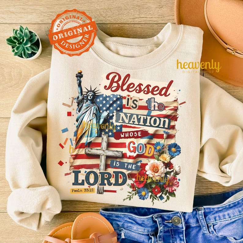 Puede incluir: Sudadera color crema con un dise&ntilde;o gr&aacute;fico que presenta la Estatua de la Libertad, una bandera estadounidense, una cruz y detalles florales. El texto dice "Blessed is the Nation Whose God is the Lord" con Salmo 33:12. Un logotipo circular "Original Designer" es visible.