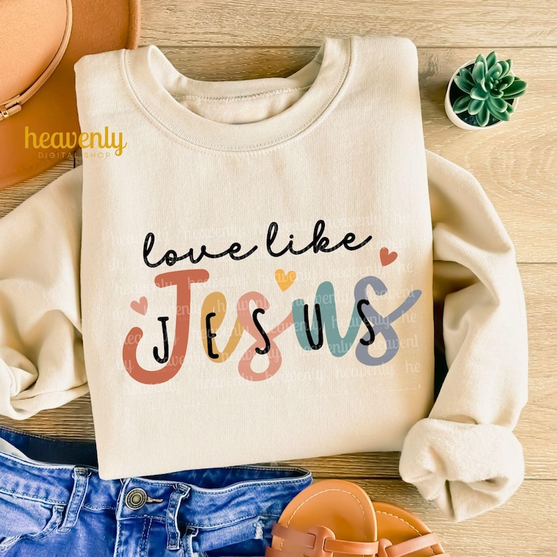 May include: Cream-colored sweatshirt with the text "love like Jesus" in black script. The word "Jesus" is in colorful block letters. A small succulent plant in a white pot is visible in the upper right corner.